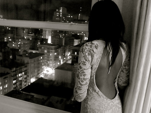 Девушка с темными волосами смотрит в окно на ночной город в белом платье с голой спиной