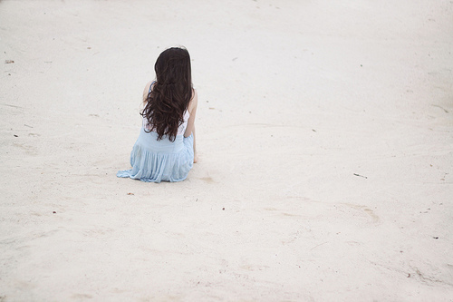 Брюнетка в голубом платье со спины сидит на песке