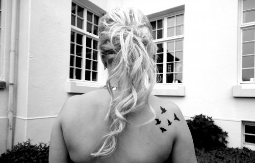 Блондинка с татуировкой птиц на плече стоит спиной во дворе дома
