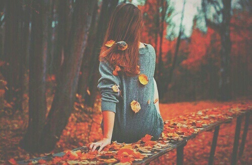 рыжая девушка в сером свитере с листьями со спины вдалеке в осенеем лесу