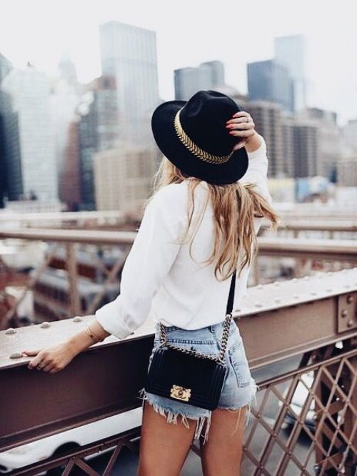 девушка со спины в шляпе и джинсовых шортах с маленькой сумочкой на фоне города