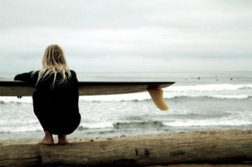 дівчина зі спини присіла на корча з дошкою для серфінгу і дивиться на океан