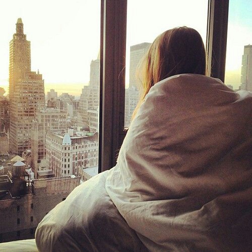 девушка укуталась в одеяло спиной без лица смотрит на город в окне