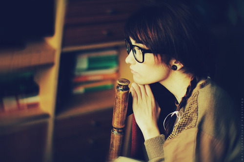 дівчина в окулярах замислилася сидячи на стільці вдома біля книжкової шафи