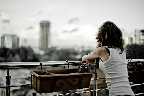  дівчина в білій майці зі спини стоїть на балконі в похмуру погоду