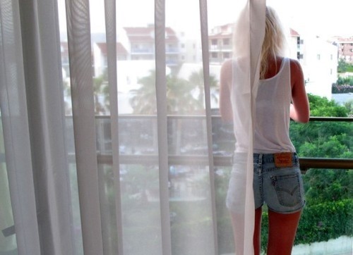 блондинка в белой майке и коротких джинсовых шортах спиной на балконе за занавеской