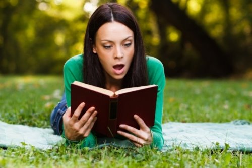 девушка с удивлением читает книгу лёжа на лужайке