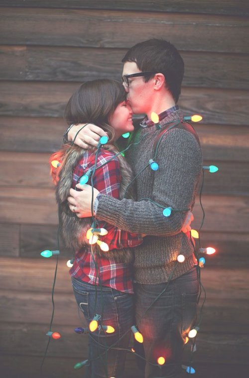 девушка в клетчатой рубашке и парень в свитере обнимаются обмотавшись новогодней гирляндой