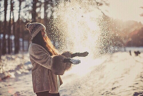 девушка с длинными волосами в шубке и шапке играет со снегом в лучах солнца