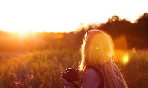 блондинка с фотоаппаратом со спины в солнечных лучах в поле