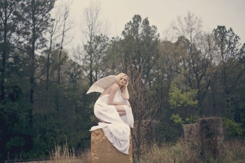 девушка в длинном белом платье с крыльями ангела сидит на пеньке