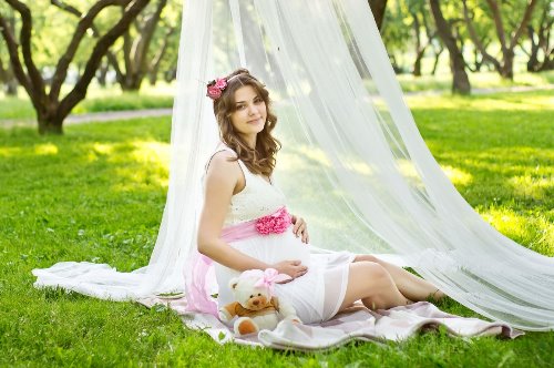 беременная в белом платье с плюшевым мишкой под деревом на траве