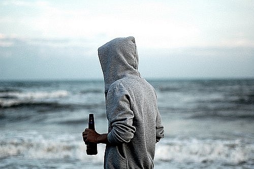 парень гуляет вдоль моря с бутылкой пива в сером реглане с капюшоном