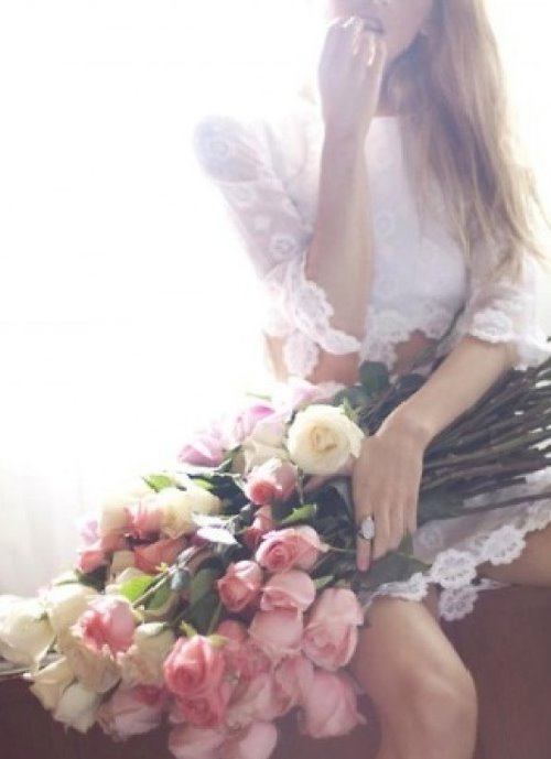 блондинка в нежном белом платье сидит на подоконнике с букетом роз на коленях