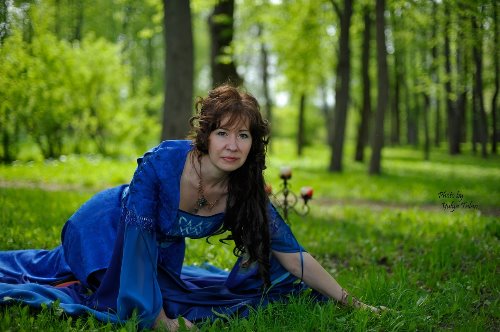 женщина в образе лесной нимфы в синем платье стоит на коленях на лесной поляне рядом подсвечник