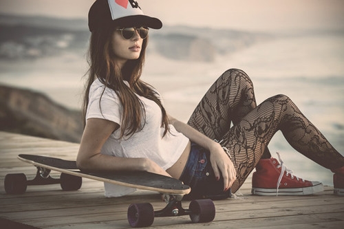 девушка в джинсовых шортах поверх колготок в очках и кепке сидит облокотившись о скейт
