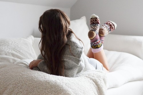 девушка в свитере и теплых носках лежит на кровати на животе прикрываясь волосами