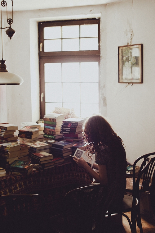 девушка со спины читает книгу среди стопок книг в комнате с высокими потолками