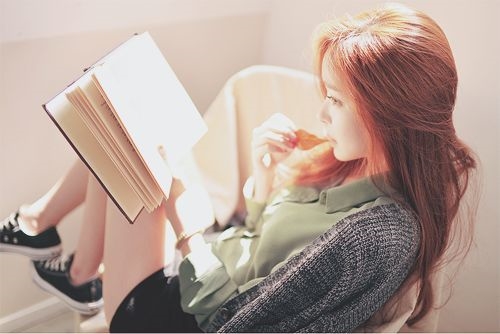 рыженькая девушка в кедах читает книгу на кресле