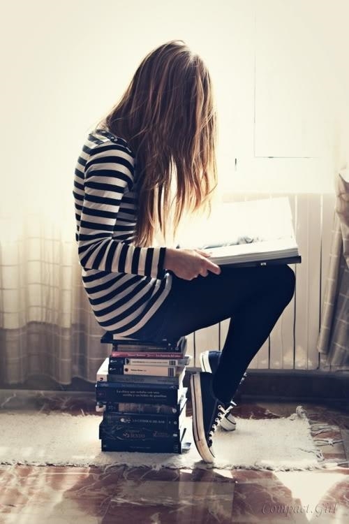 девушка в кедах и полосатой кофте сидит на стопке книг и читает