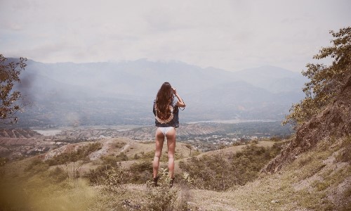 девушка в белых трусиках с длинными волосами со спины в горах смотрит в бинокль