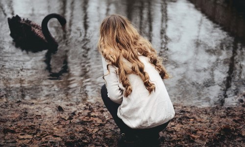 девушка с светлыми локонами со спины присела у водоёма кормить черного лебедя весной