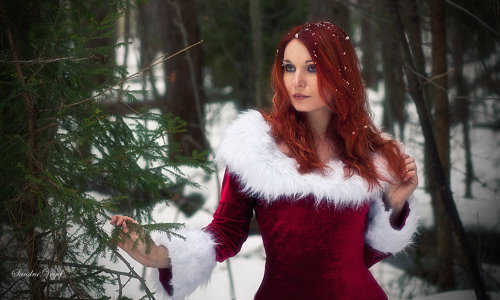 рыжая девушка в зимнем лесу возле ели с снегом на волосах в красном новогоднем платье