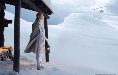 девушка в белом с подсвечником в руках смотрит вдаль на зимние просторы позади нее стопка дров и дом