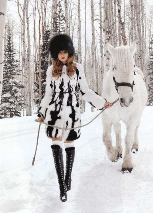 девушка с вьющимися светлыми волосами в меховой шапке ведет по зимнему лесу белую лошадь