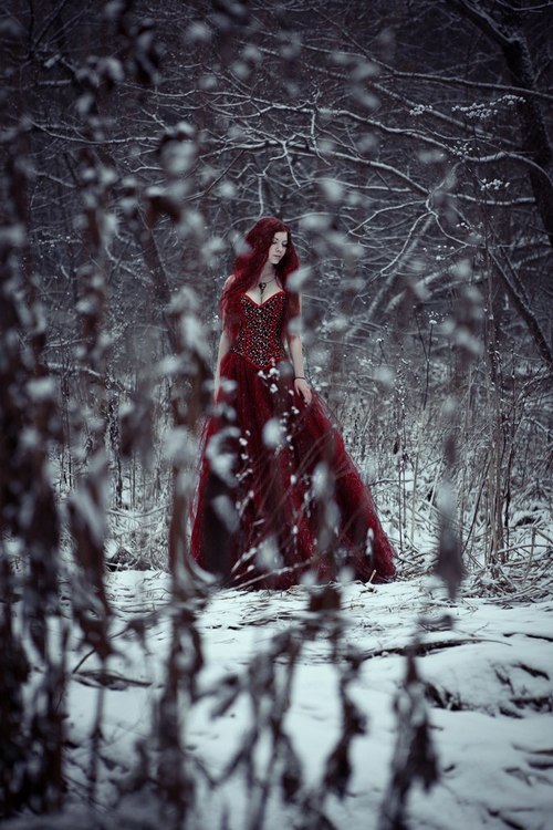 девушка с красными волосами в бордовом длинном бальном платье среди деревьев зимой в снегу