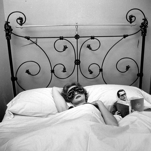 большая женщина спит по центру кровати, а рядом с ней лежит маленький мужчина в очках, который читает книгу