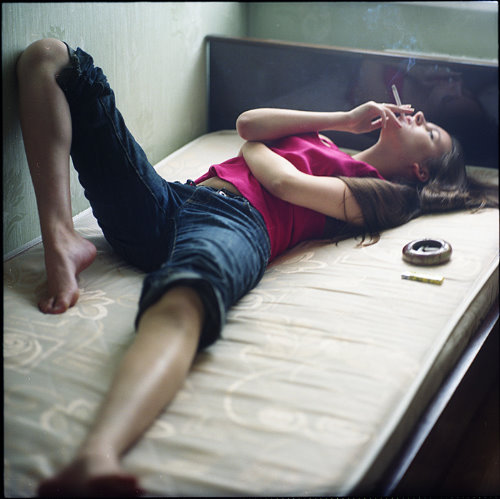 русая девушка лежит на кровати без постельного белья с сигаретой и смотрит в потолок одета в розовую футболку и синие джинсовые шорты
