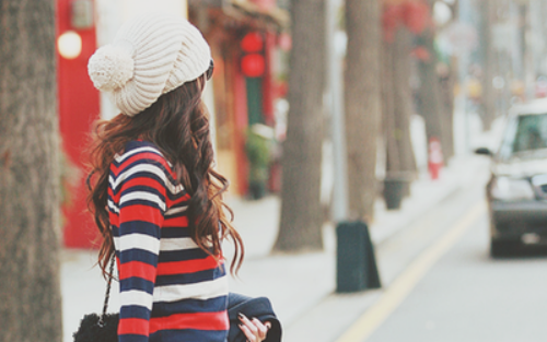 девушка в полосатом свитере переходит через дорогу в белой шапке с помпоном и локонами коричневых волос