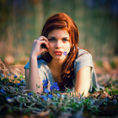 пронзительный взгляд синеглазой девушки с рыжими волосами которая лежит на цветочной поляне и держит губами фиолетовый цветок
