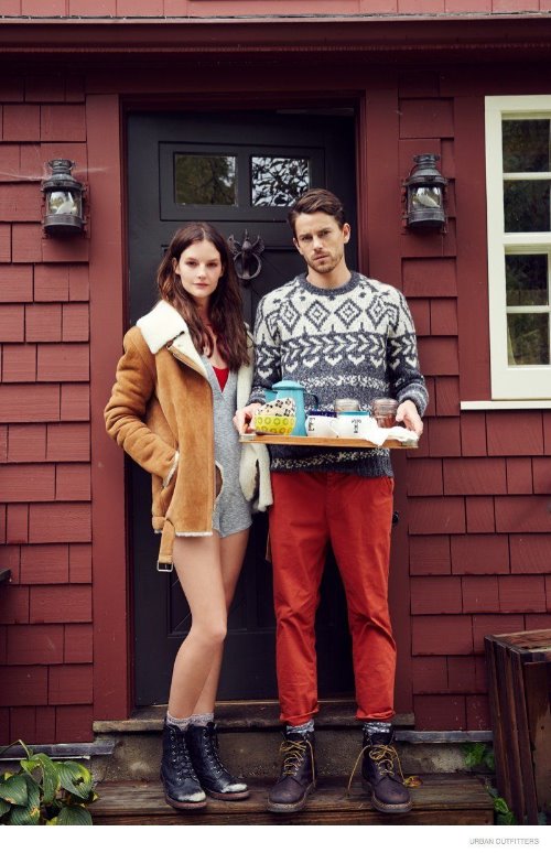 красивая пара с коричневыми волосами стоит на крыльце дома с подносом с завтраком она в коротком кожухе, он в вязаном сером свитере и красных брюках