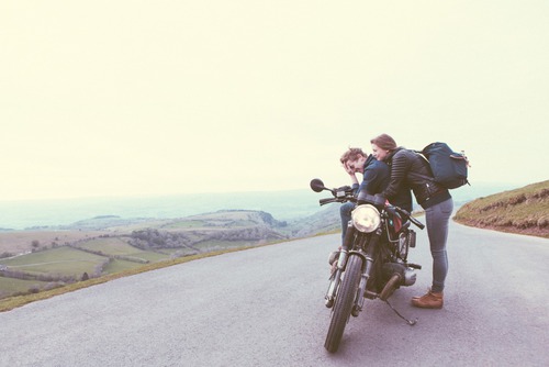Парень смеется сидя на мотоцикле, а девушка с рюкзаком обнимает его со спины.