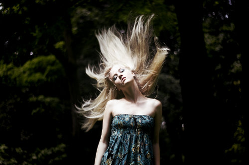 блондинка летом на размытом фоне деревьев трясет волосами в синем платье