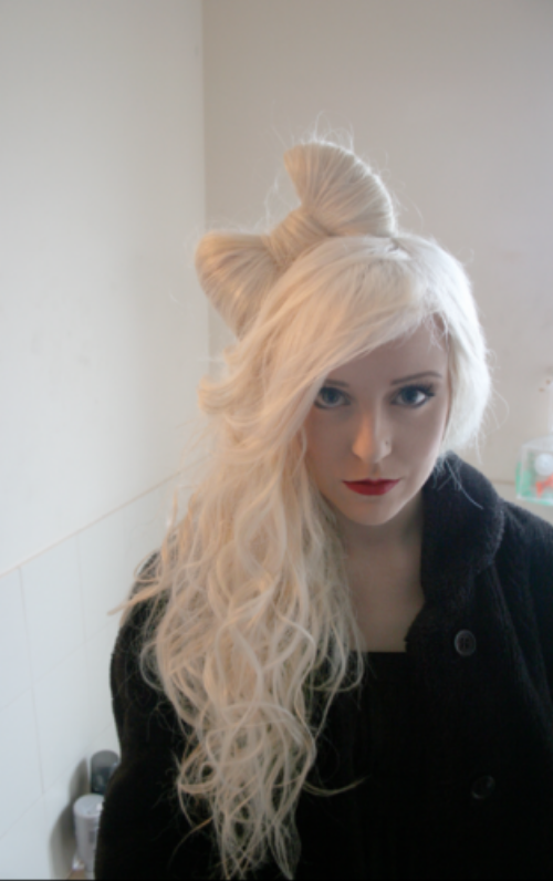 длинноволосая блондинка с прической бант из волос с ярко накрашенными красными губами и бледной кожей стоит в пальто в комнате