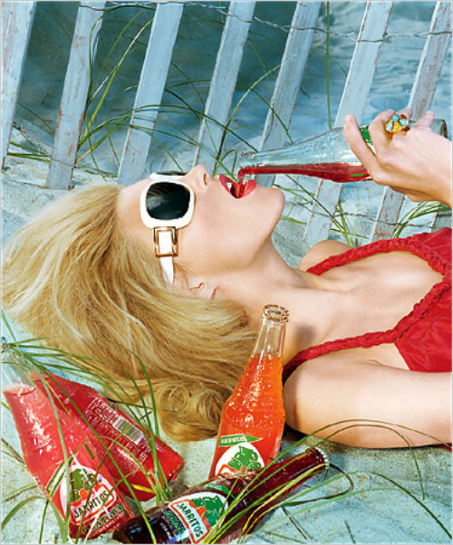 блондинка в красной майке солнечных очках лежит у забора в траве среди бутылок и пьет лимонад
