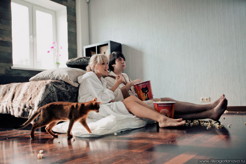 блондинка в белом халате с челкой ест попкорн со своим парнем брюнетом в спальне смотрят кино упершись об кровать рядом проходит кошка коричневого цвета