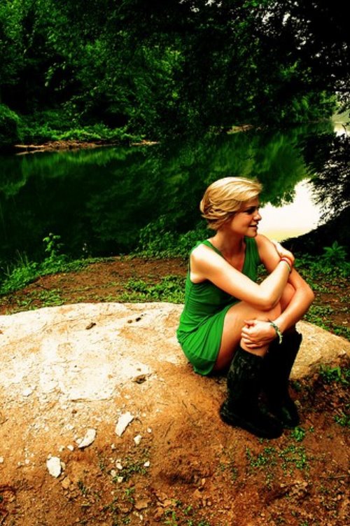 блондинка с короткими волосами в зеленом платье и резиновых сапогах сидит на берегу реки на камне