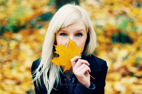 блондинка с голубыми глазами и ногтями держит желтый кленовый лист прикрывая им лицо на размытом осеннем фоне