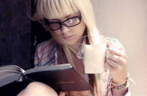 Блондинка с прямой челкой в клетчатой рубашке в очках с книгой и кружкой в руках. 