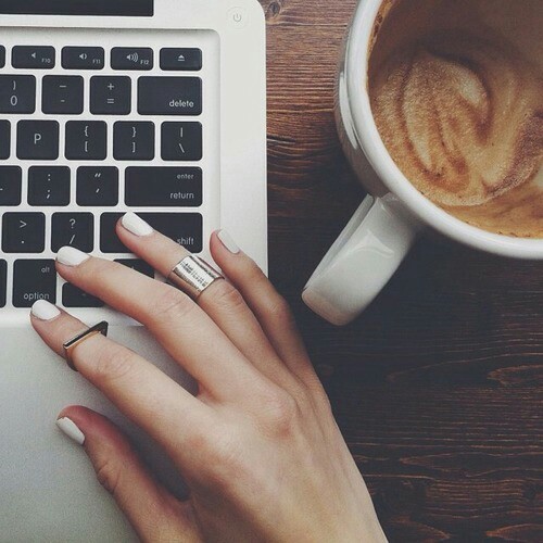 Девушка с капучино работает за ноутбуком с белыми ногтями и двумя фаланговыми кольцами на указательном и безымянном пальцах