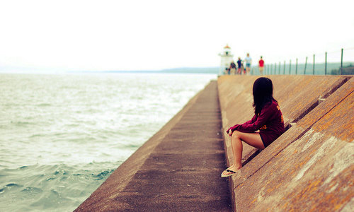Девушка без лица сидит у воды смотрит на маяк вдалеке