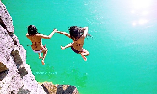 две девушки прыгают со скалы в море держась за руки спиной