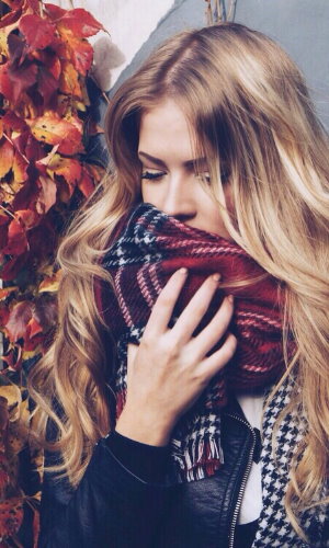 Девушка с длинными светлыми вьющимися волосами кутается в клетчатый красный шарф осенью.