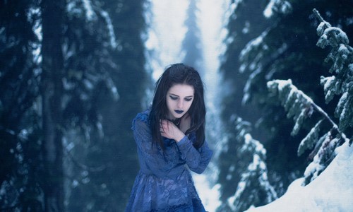Девушка брюнетка в синем платье с синими губами замерзла зимой в лесу среди заснеженных елей