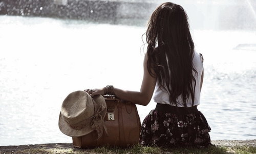 Девушка с сумкой и шляпой со спины сидит у воды