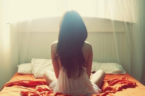 Девушка с густыми волосами сидит на кровати в позе лотоса спиной
